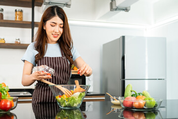 Happy Asian woman wearing apron, adding cherry tomato while tossing the vegetarian salad with wooden spatulas. Preparing a healthy salad with fresh vegetables in the home kitchen.