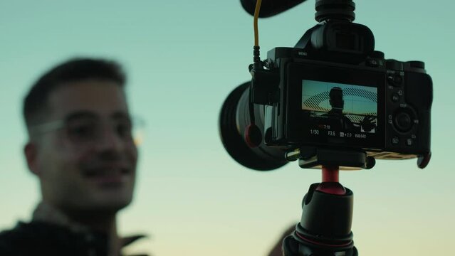 Happy, good-looking male content creator talking to the camera during sunset. Slow motion of a young, handsome influencer guy vlogging with a camera during sunrise. Close-up with focus on the camera.