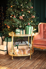 Decorative golden trolley table with gifts and decor. Decorated Xmas tree with lights glowing garlands. Stylish loft living room interior with pink sofa. Christmas at home. New year winter home decor.