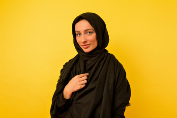Elegant stylish woman with blue eyes and charming smile wearing black burqa posing over yellow wall