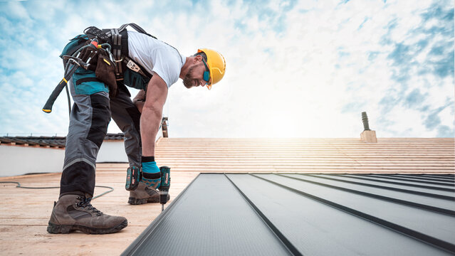 A roofer with a safety harness and tool belt is working with a electric screwdriver on the roof. He is anchoring the metal roofing with a screws.