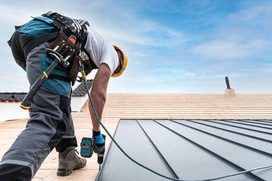 A roofer with a safety harness and tool belt is working with a electric screwdriver on the roof. He is anchoring the metal roofing with a screws.