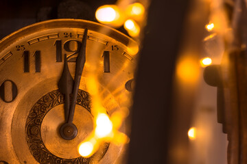 Close-up of the golden dial of an old clock pointing to midnight, illuminated by the warm light of Christmas lights.