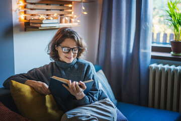 Young smiling cheerful woman in a warm sweater and eyeglasses reading a book while sitting on the couch in the room - obrazy, fototapety, plakaty