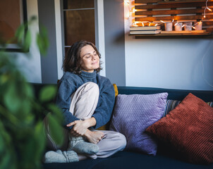 Young adult happy cheerful woman in warm sweater sitting on sofa enjoying resting and relaxing with...