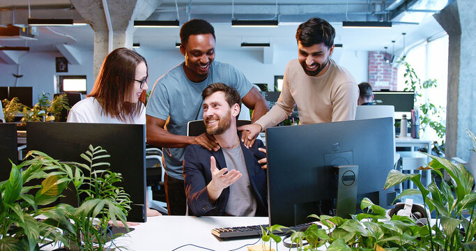 Multiethnic team of males and female cooperating in coworking space. IT team of developers working, laughing and talking in office. Employees of programming company. Brainstorming meeting.