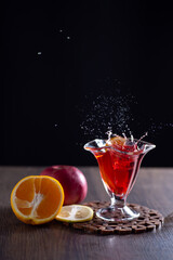 There is sangria in a glass on the table. Splashes from falling ice. Nearby lie a tangerine, a lemon and an apple.