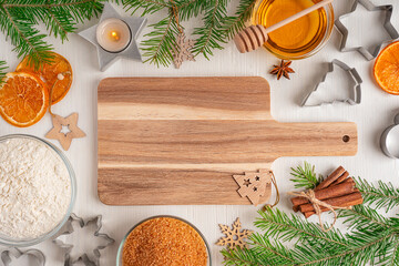Top view of ingredients for cooking of cake or biscuit with wooden copping board, sugar, honey, flour, cinnamon and cookie cutters on white wooden table with burning candle, dried oranges and fir tree