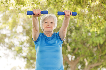 Donna anziana con i capelli grigi e un maglietta sportiva celeste fa esercizio fisico con dei pesi in un parco.