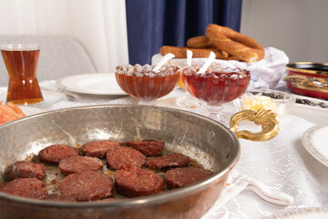 Sausage called sucuk, selective focus close up photo of fried sausage called sucuk in pan. Traditional Turkish breakfast table. Glass of tea, jar, bagel called simit. Family Sunday morning routine.