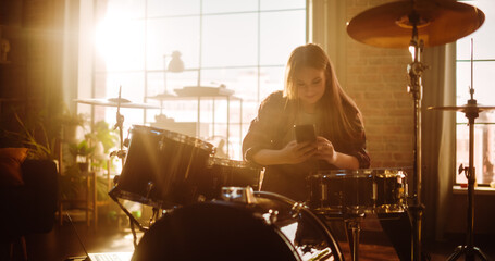 Fototapeta na wymiar Young Female Pulls out Her Smartphone to Answer a Social Media Message. Expressive Drummer Finished Playing Drums in a Loft Music Rehearsal Studio Filled with Light. Music Artist Learned a Drum Solo.