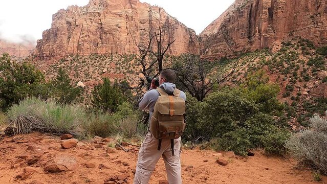 A photography wearing a backpack walks into the video and begins taking photographs in Zion National Park in Utah.