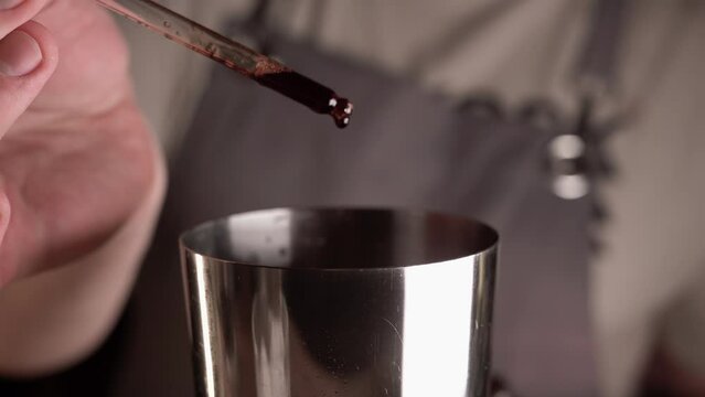Bartender is making a cocktail at bar counter, adding some bitter liquid with pipette in the shaker