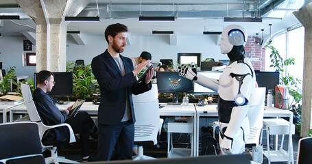 Man programming robot with tablet computer in hands in office. Male developer checking work of robotic by device. Coworking space. Humanoid moving and executing instructions and orders.