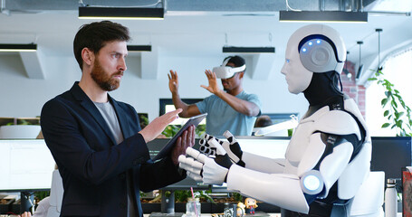 Man programming robot with tablet computer in hands in office. Male developer checking work of...