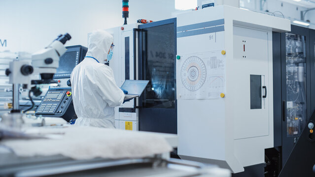 Heavy Industry Manufacturing Factory: Engineer in Sterile Coverall Working on Laptop Computer, Examining Industrial CNC Machine Setting and Configuring Functionality. - Powered by Adobe
