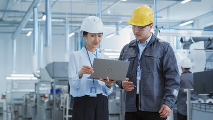 Portrait of Two Young Asian Heavy Industry Engineers in Hard Hats Standing with Laptop Computer and Discussing Work Process in a Factory. Two Manufacturing Employees Chatting in Production Facility.