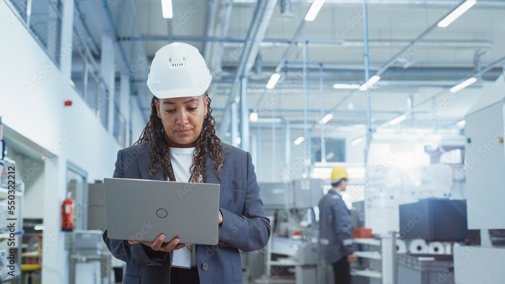Poster portrait of an african female engineer in hard hat standing and using laptop computer at electronic 