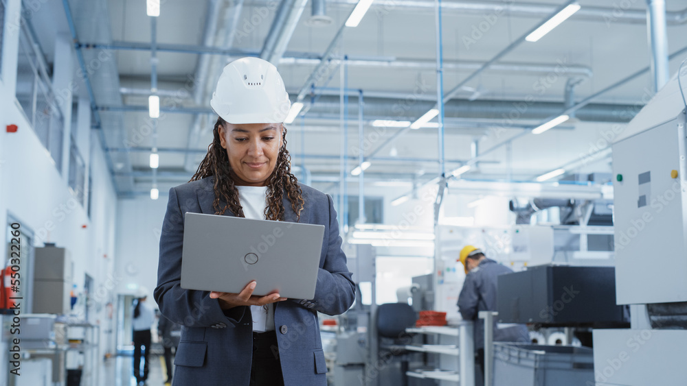 Fototapete portrait of a happy black female engineer in hard hat standing and using laptop computer at electron