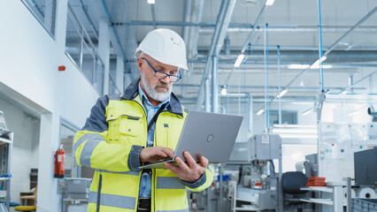 Portrait of a Bearded Middle Aged Engineer Standing in a Factory Facility, Wearing a High Visibility Jacket and a White Hard Hat. Heavy Industry Specialist Working on Laptop Computer.