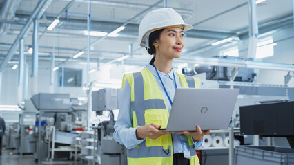 A Happy and Smiling Asian Female Engineer in White Hard Hat Standing with Laptop Computer at Electronic Manufacturing Factory. Successful Employee Focusing on PC Screen