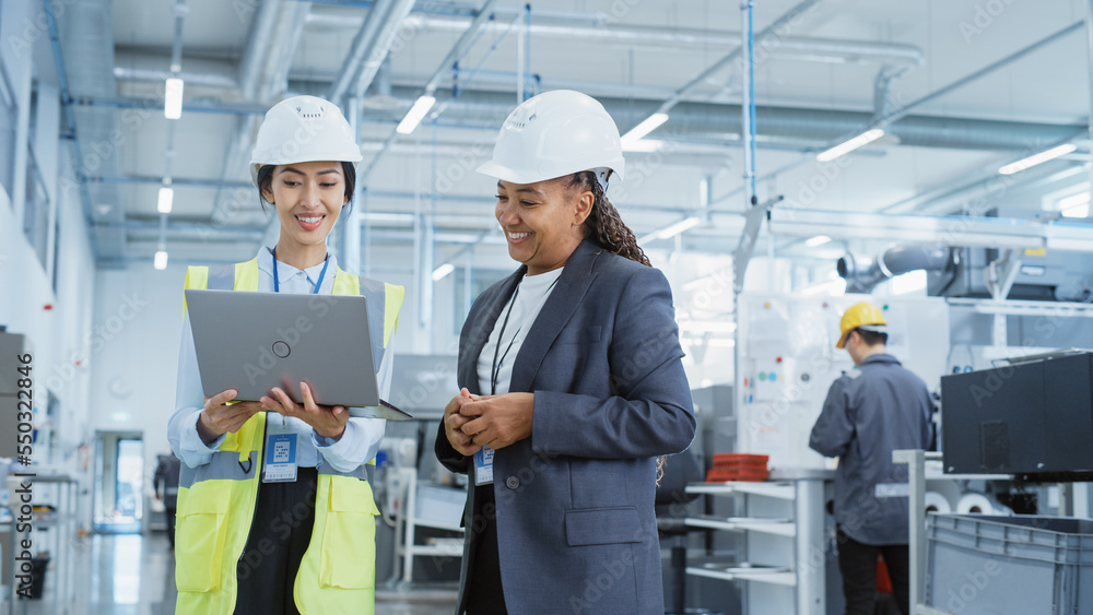Poster portrait of two female employees in hard hats at factory discussing assignments at industrial machin