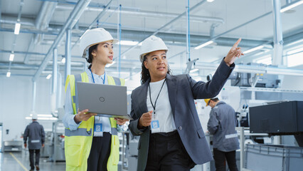 Portrait of Two Female Employees in Hard Hats at Factory. Discussing Job Assignments at Industrial Machine Facility, Using Laptop Computer. Asian Engineer and African American Technician at Work.