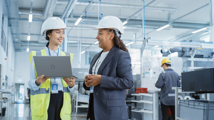 Portrait of Two Female Employees in Hard Hats at Factory Discussing Assignments at Industrial Machine Facility, Using Laptop Computer. Smiling Asian Engineer and African American Technician at Work.