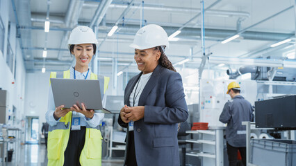 Portrait of Two Female Employees in Hard Hats at Factory Discussing Assignments at Industrial Machine Facility, Using Laptop Computer. Smiling Asian Engineer and African American Technician at Work.