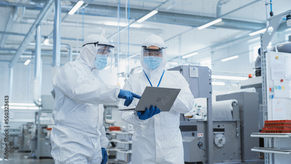 Poster two scientists walking in a heavy industry factory in sterile coveralls and face masks, using laptop