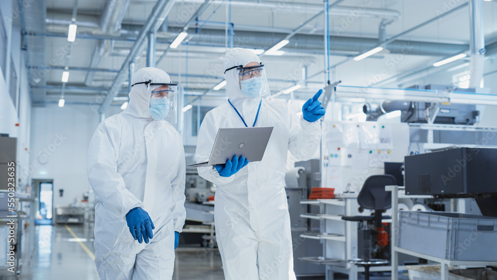 Poster two scientists walking in a heavy industry factory in sterile coveralls and face masks, using laptop