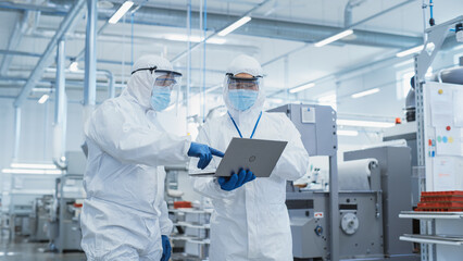 Two Scientists Walking in a Heavy Industry Factory in Sterile Coveralls and Face Masks, Using...