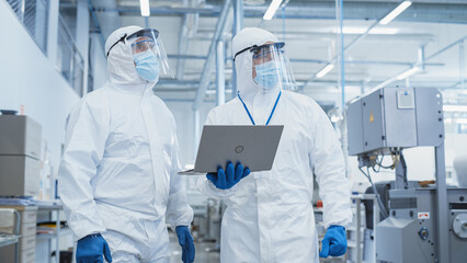 Two Scientists Walking in a Heavy Industry Factory in Sterile Coveralls and Face Masks, Using Laptop Computer. Examining Industrial Machine Settings and Configuring Production Functionality.