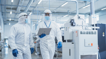 Two Scientists Walking in a Heavy Industry Factory in Sterile Coveralls and Face Masks, Using...