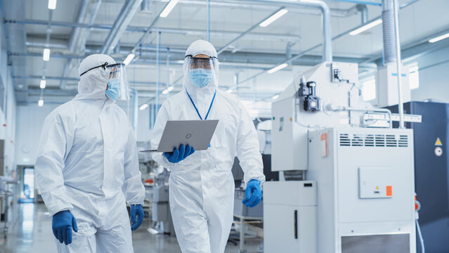 Two Scientists Walking in a Heavy Industry Factory in Sterile Coveralls and Face Masks, Using Laptop Computer. Examining Industrial Machine Settings and Configuring Production Functionality.