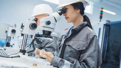 Young Asian Industrial Scientist and Older Engineer Working at a Desk in a Factory Facility, Using Microscopes to Inspect the Manufacturing Production Parts for Quality.
