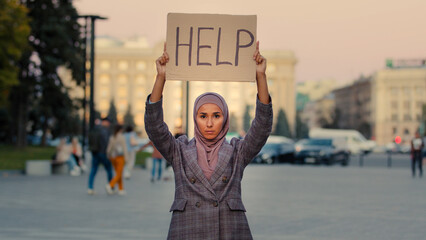 Sad lonely poor muslim girl wears hijab woman stands in city looking at camera demonstrates shows cardboard banner text help needs protection work, unemployment discrimination problem concept