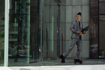 young businessman in roller skates and grey suit holding coffee to go and laptop near building with glass doors.