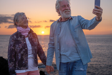 Smiling senior couple standing in the sunset light on the rocks taking a selfie with the orange sun...