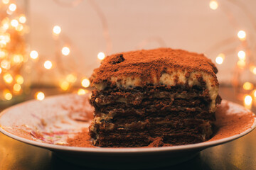 A piece of tiramisu sprinkled with cocoa on a white plate and yellow Christmas lights. Delicious sweet dessert. Homemade cake. Holiday atmosphere