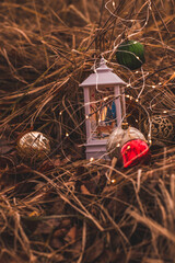 Beautiful New Year background. Christmas atmosphere. Golden, red and green Christmas ornaments, white lamp and Christmas lights in the dry grass. Garden decoration in winter holidays