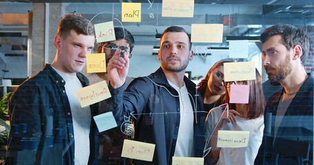 Diverse people of startup project standing in front of board with tasks and to-do notes, drawing...
