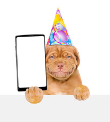 Smiling mastiff puppy wearing party cap holds big smartphone with white blank screen in it paw, showing close to camera above white banner. Empty free space for mock up, banner