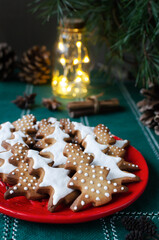 Festive cookies decorated with white icing on a red plate on a green background with lights. Homemade ginger and cinnamon Christmas cookies. Vertical orientation. Selective focus.
