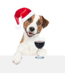 Happy Jack russell terrier puppy wearing red santa hat holds glass of red wine and looks above  empty white banner. isolated on white background