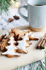Festive cookies decorated with white icing on a wooden cutting board with a hot drink in a blue cup on a marble background with lights. Homemade ginger and cinnamon Christmas cookies. 