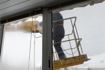 Worker man in overalls in the crane basket remove snow and ice from city house balcony roof at winter day. Roof cleaning, utility service, safety works