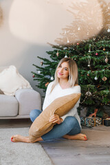 Pretty woman in jeans and a white sweater is sitting on the sofa against the background of a Christmas tree in the living room and waiting for the holiday