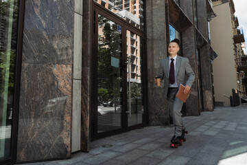 smiling man in suit and roller skates riding with coffee to go along building on urban street.