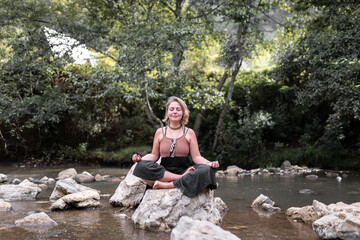 A young blonde woman is meditating sitting on a river rock